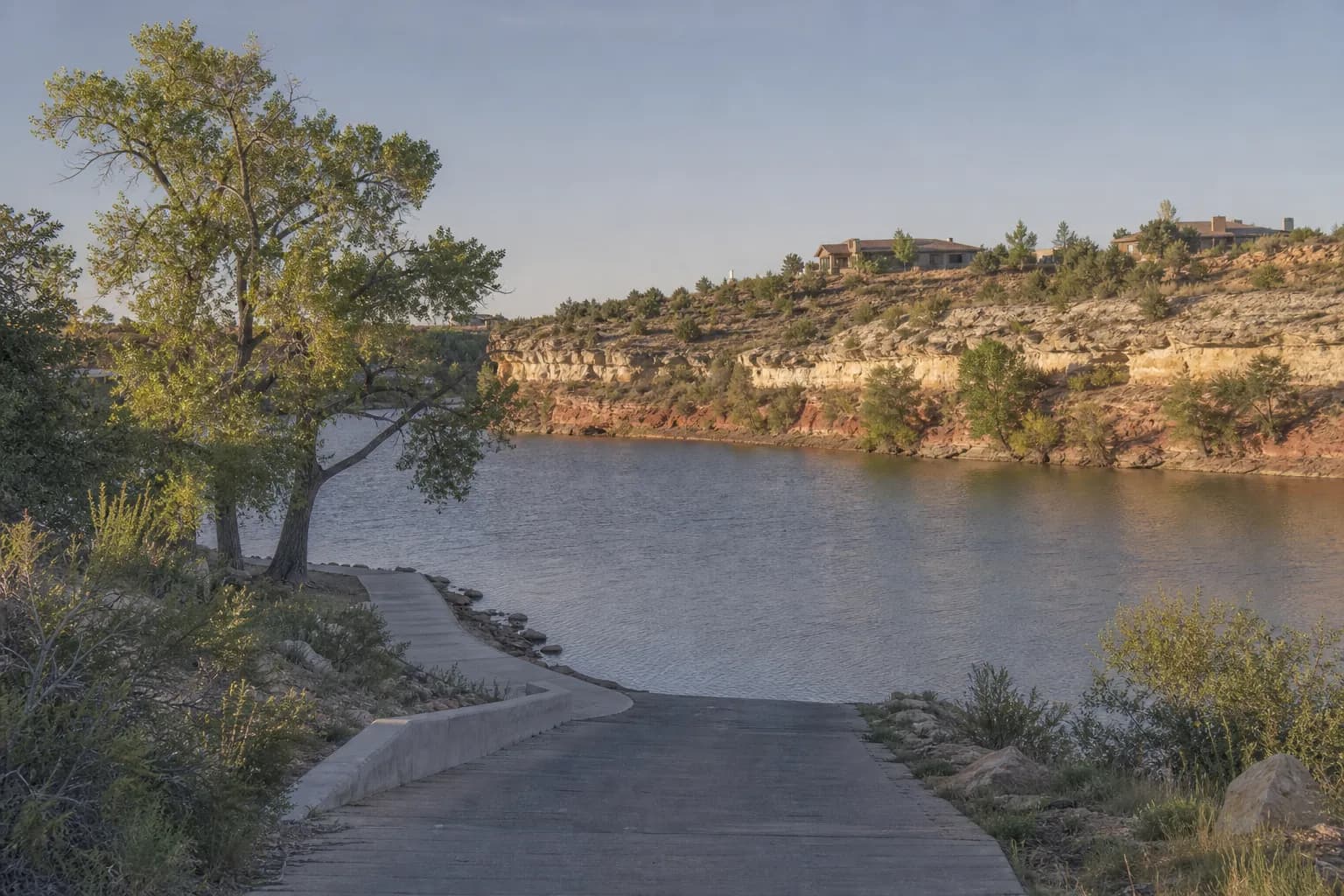 Small canyon lake with homes on the rim, layered caliche and red rock walls, cottonwoods and a concrete walkway along the shore