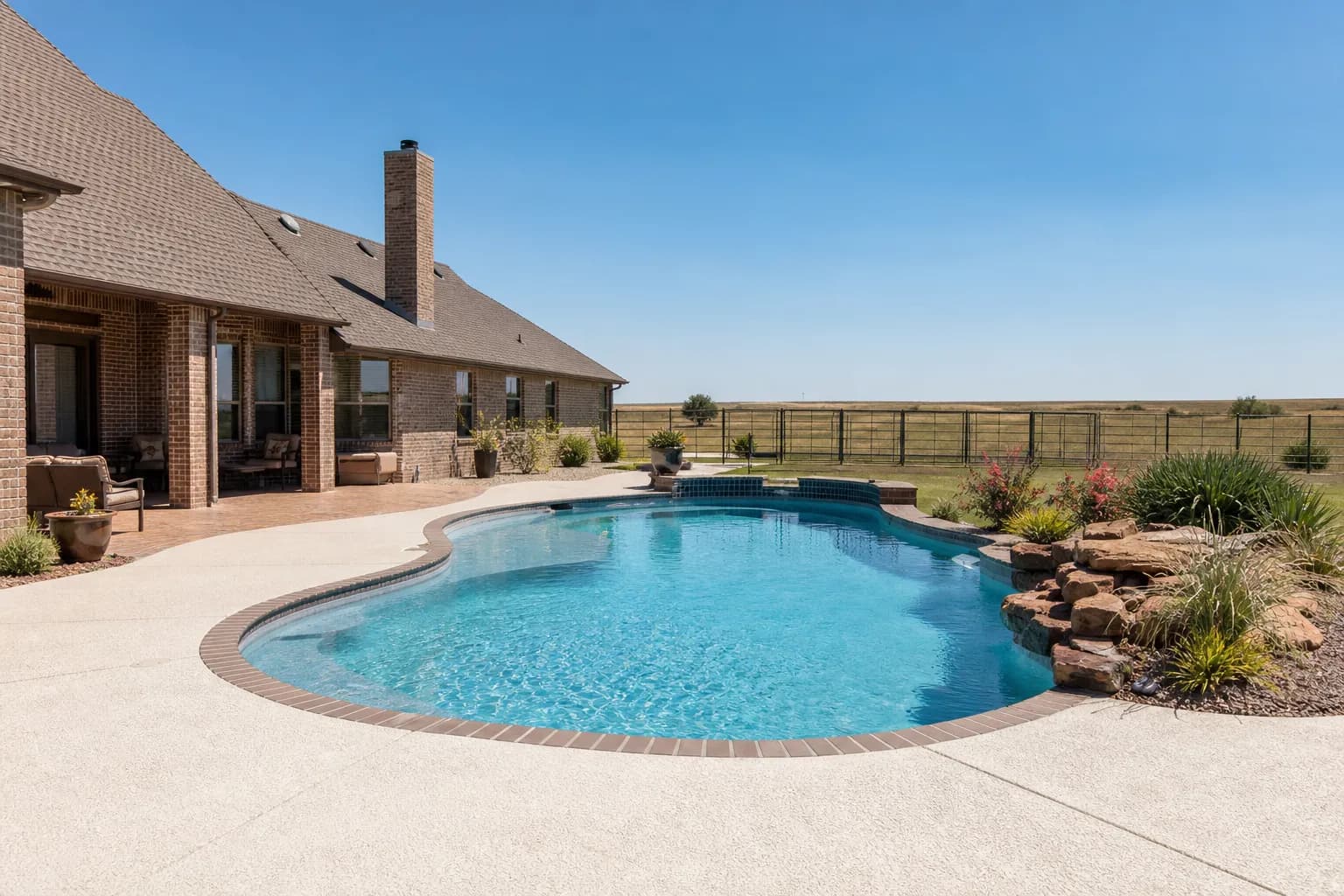Backyard swimming pool with a light-colored textured concrete deck beside a brick home under a clear sky