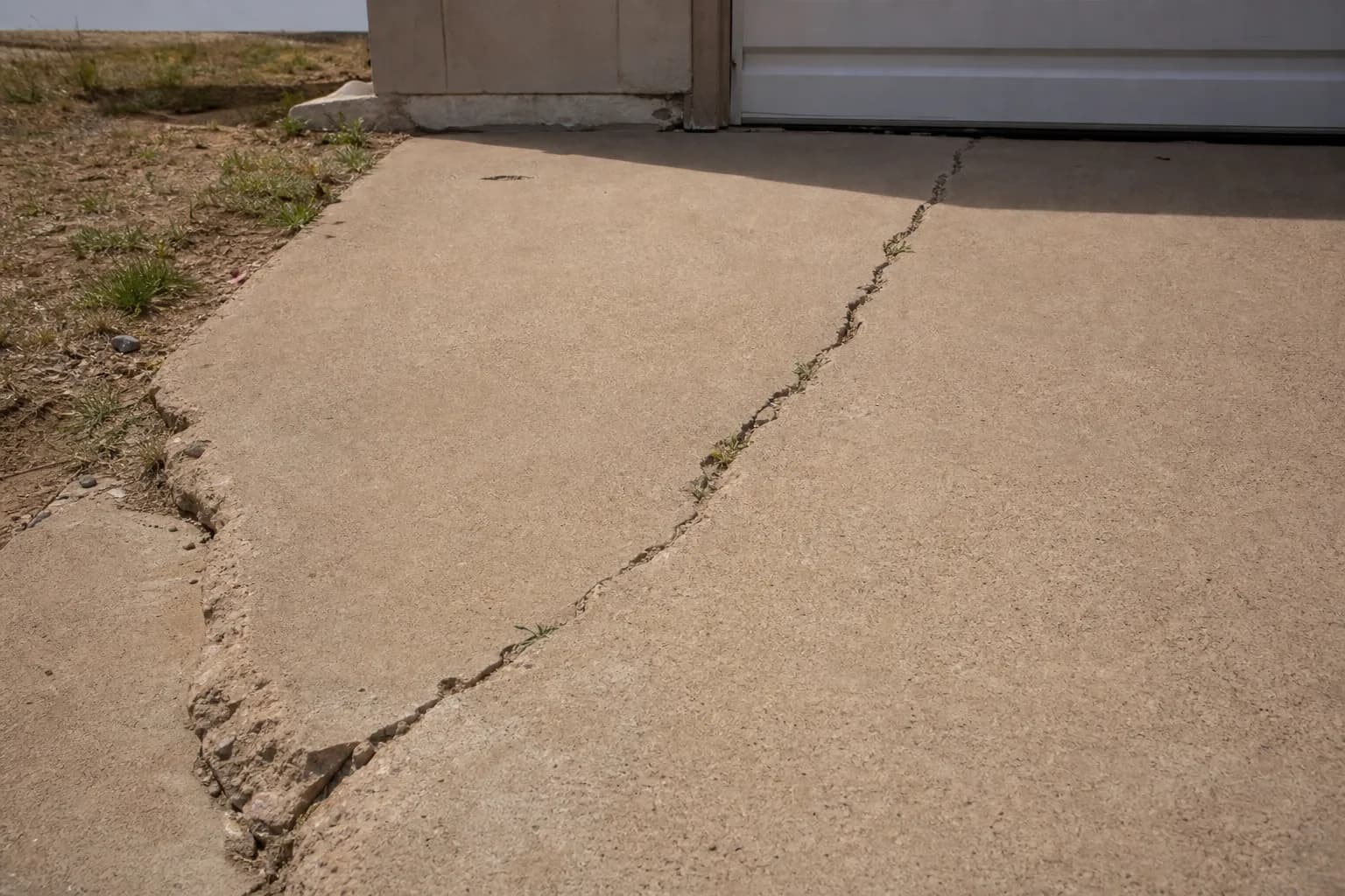 Older concrete driveway with a long diagonal crack and a chipped edge near a garage, with weeds growing in the crack
