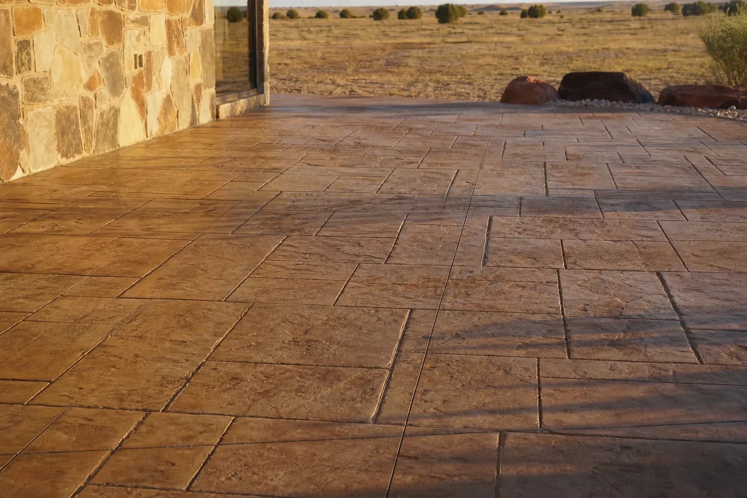 Stamped concrete patio in a large stone pattern in warm amber and brown tones beside a stone wall, with open plains beyond