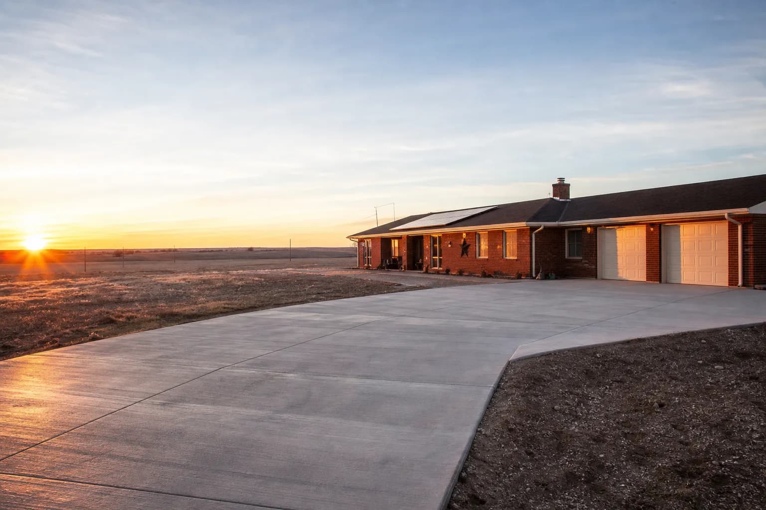 New broom-finish concrete driveway leading to a red brick ranch house on open West Texas land at sunset, with a flat horizon and wide sky