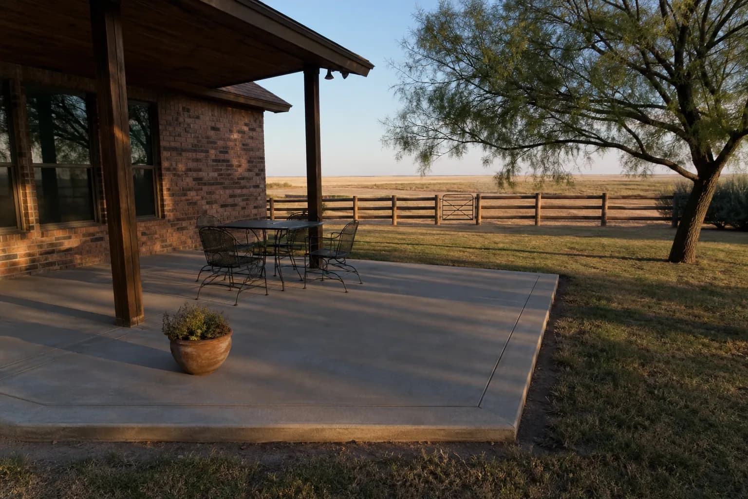 Covered concrete patio behind a brick home, with a mesquite tree and a flat horizon beyond the fence